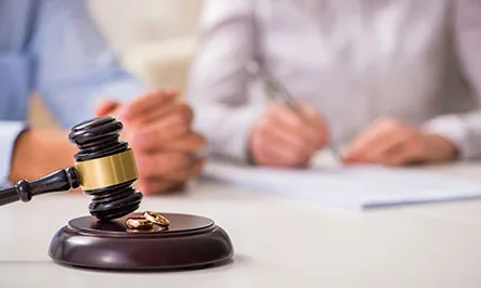 A gavel and wedding rings on a table with two people in the background, one writing on papers, suggesting a legal or divorce proceeding.