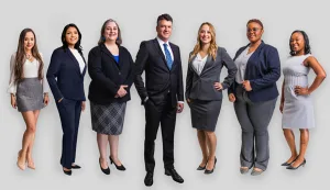 Seven professionally dressed adults, six women and one man, stand in a row against a plain light background, facing forward and smiling.