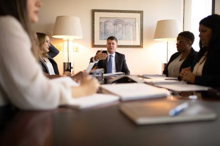 Five people in business attire have a meeting around a conference table with notepads, laptops, and documents; one man speaks while others listen.