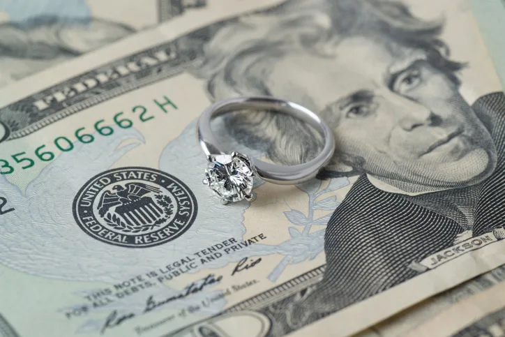 A diamond engagement ring rests on top of U.S. twenty-dollar bills.