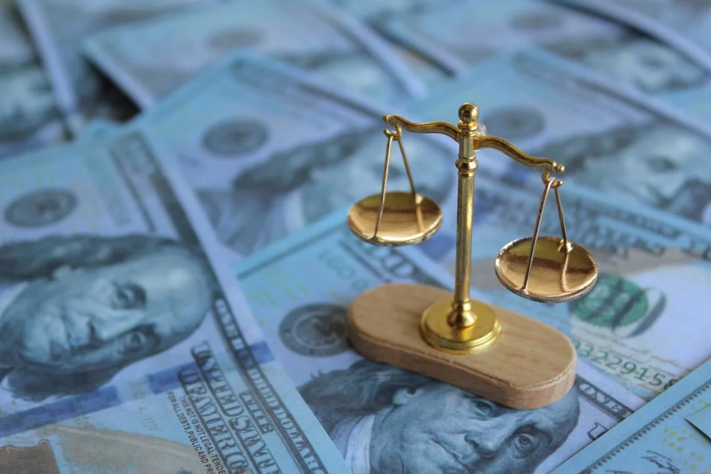 A small golden balance scale sits on a pile of US hundred dollar bills, symbolizing the relationship between money and justice.