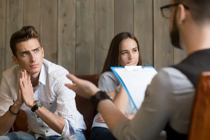 A man and a woman sit on a couch, listening intently to a person holding a clipboard who is speaking to them.
