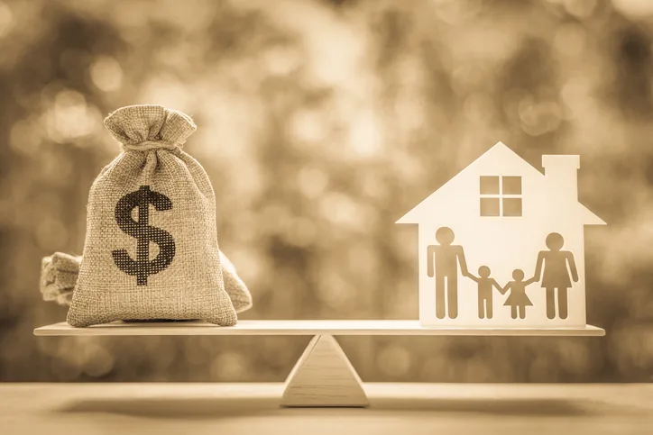 A sack with a dollar sign and a cutout of a family in a house are balanced on a seesaw, symbolizing financial balance and family.