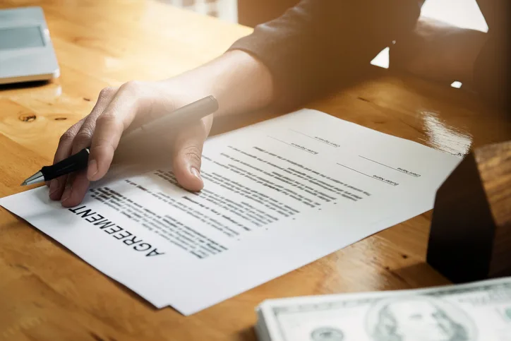 A hand holding a pen is poised over an agreement document on a wooden desk, with cash and a laptop partially visible nearby.