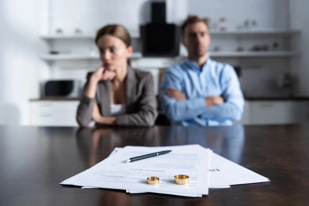 A man and woman sit apart at a table with divorce papers, a pen, and two wedding rings in focus on the table in front of them.