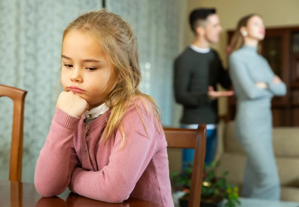 A young girl sits at a table looking sad while two adults argue in the background.