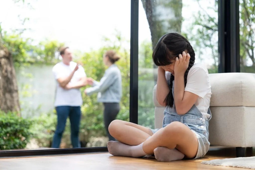 A young girl sits on the floor covering her ears, while two adults argue outside in the background, visible through a glass door.
