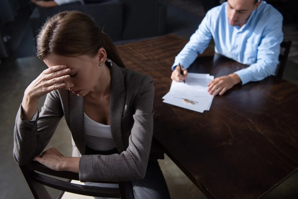 A woman sits with her hand on her forehead looking distressed while a man signs papers at a table in the background.