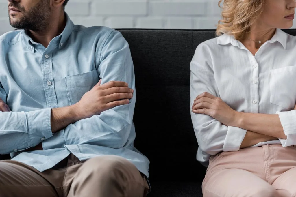 A man and woman sit on a couch with arms crossed, turned away from each other, appearing upset or in disagreement.