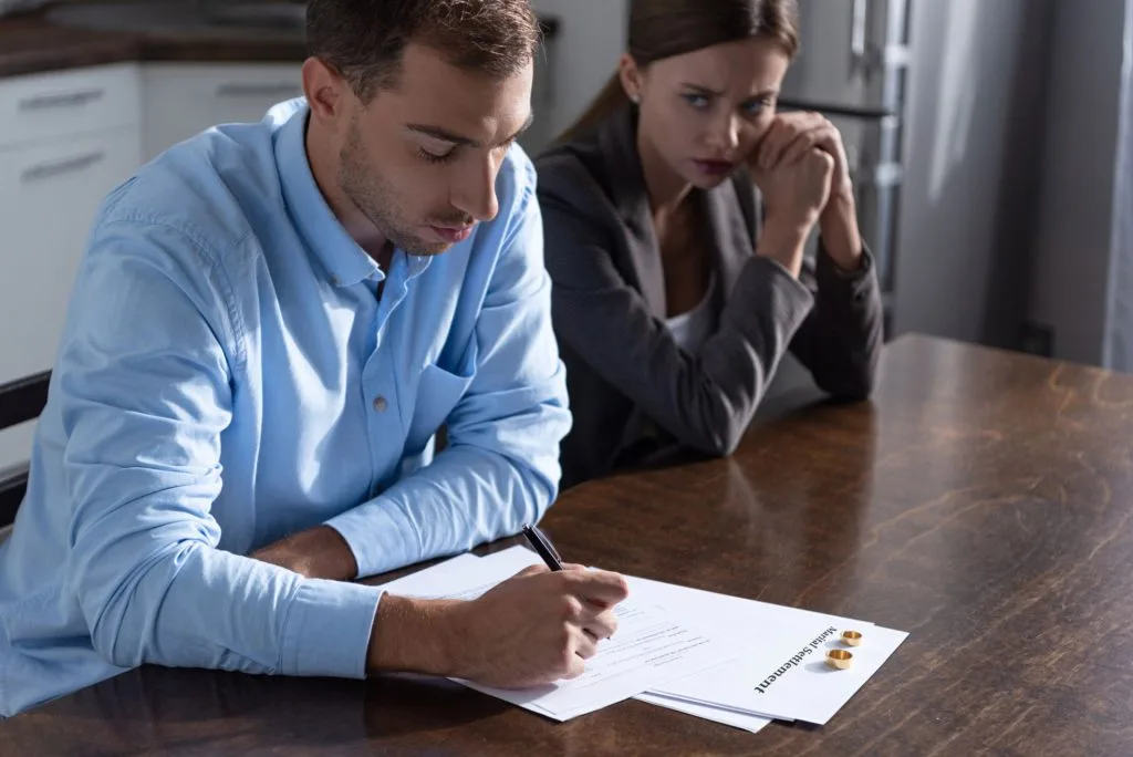 A man signs divorce papers at a table while a woman sits next to him, looking at him. Two wedding rings are placed on the documents.