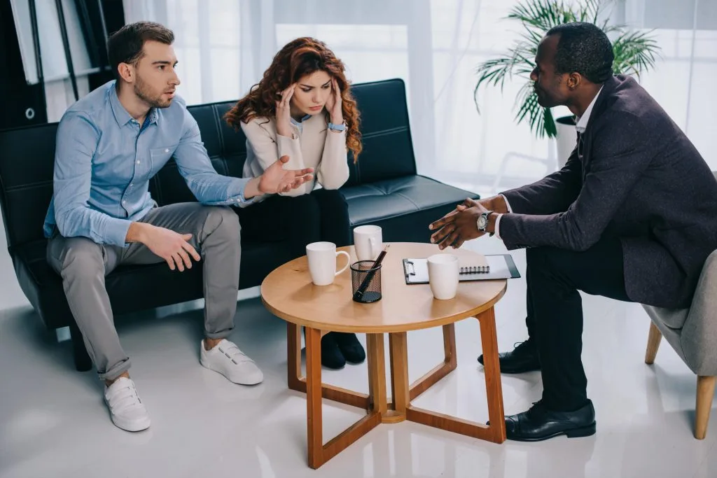 A man and woman sit on a couch talking with another man across from them in an office setting. Three coffee cups, a notebook, and an hourglass are on a small round table between them.