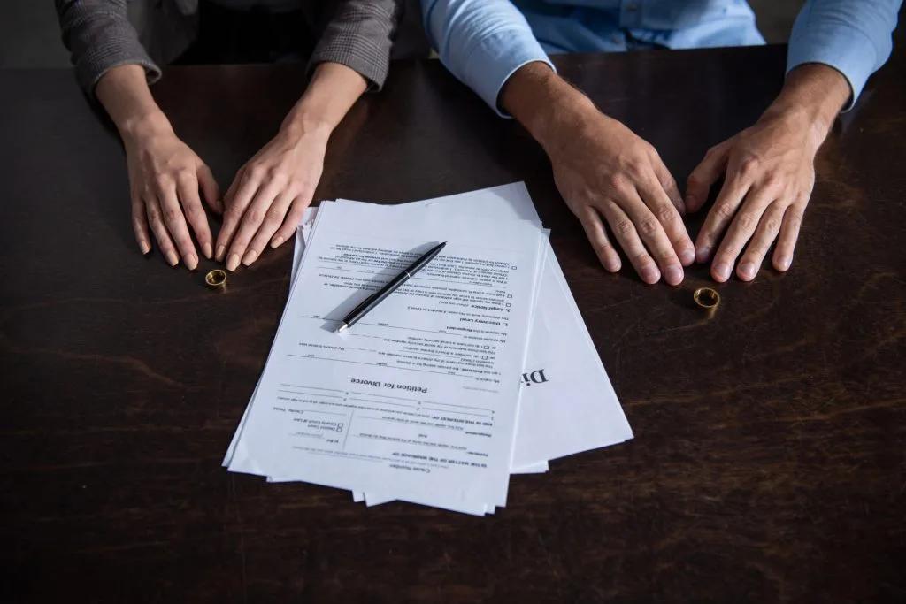 Two people sit at a table with divorce papers, a pen, and wedding rings placed separately in front of them.