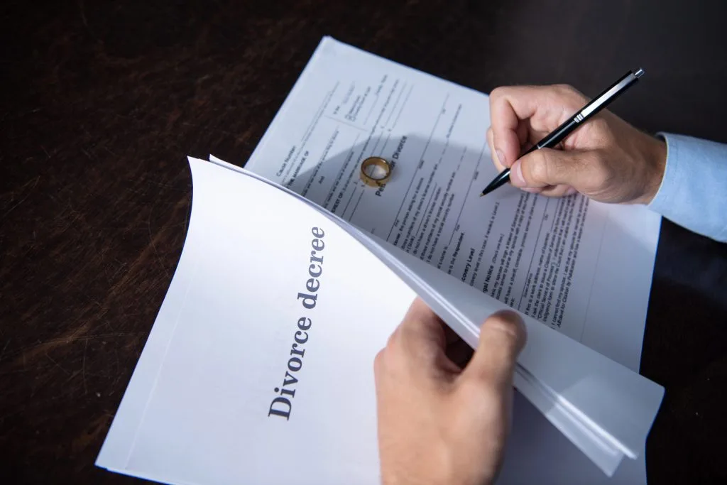 A person signs official divorce documents on a dark desk with a gold wedding ring placed on the paperwork.